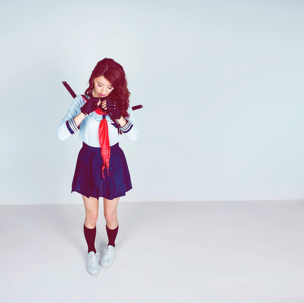 A young woman in a schoolgirl cosplay stands with swords in a studio setting.